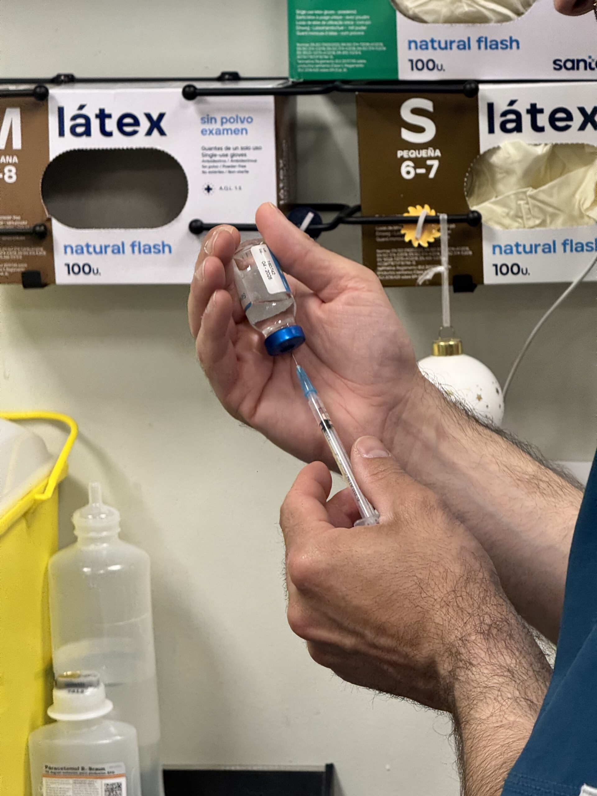 Profesional sanitario extrayendo medicación de vial con jeringa en entorno clínico con guantes de látex al fondo